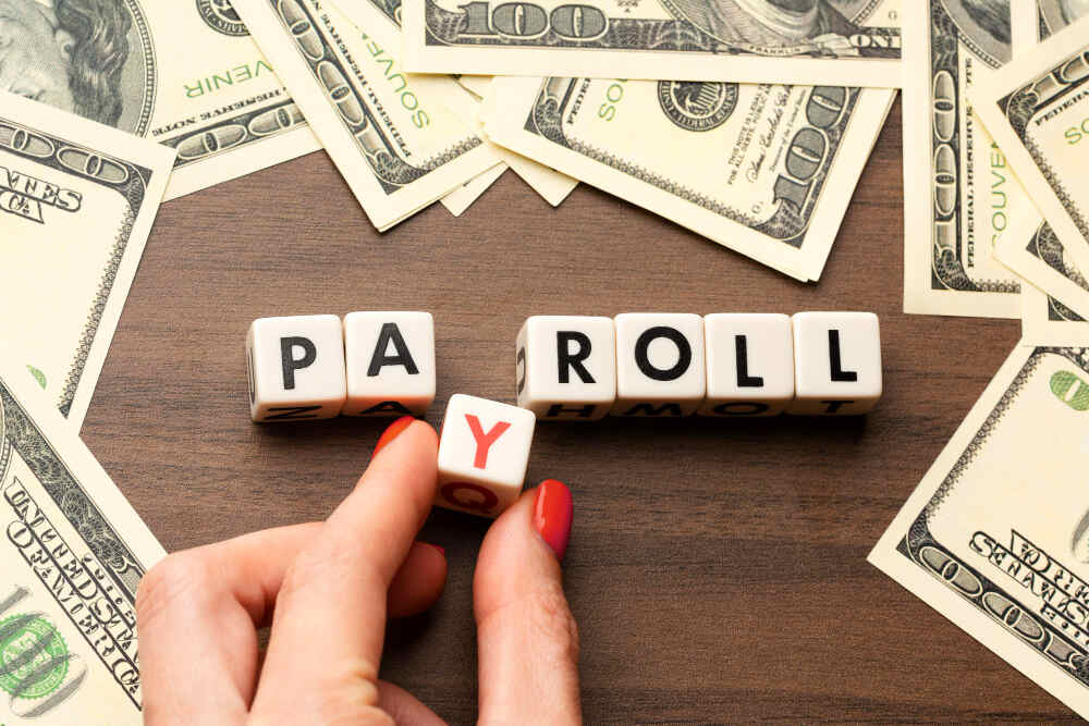 A person's hand holding six dice that spell out the word "payroll.